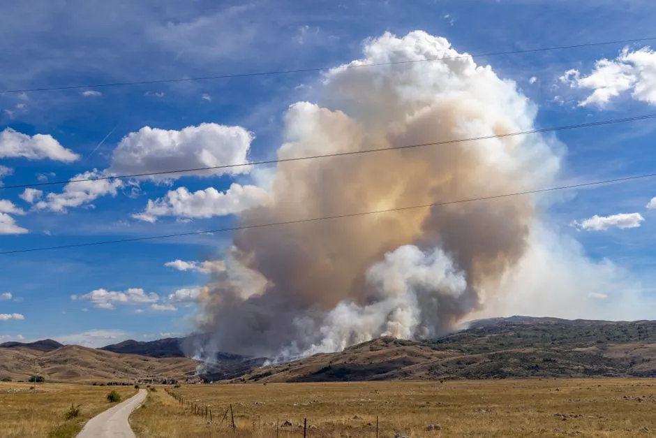 Dramatic image of a large forest fire with dense smoke in a mountainous landscape under a blue sky.